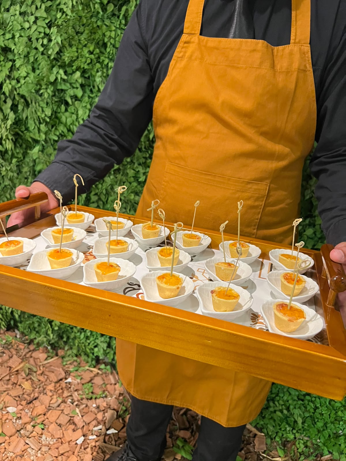 garçom de avental mostarda segurando bandeja de madeira com porções de quiche em miniaturas servidas em bowls 