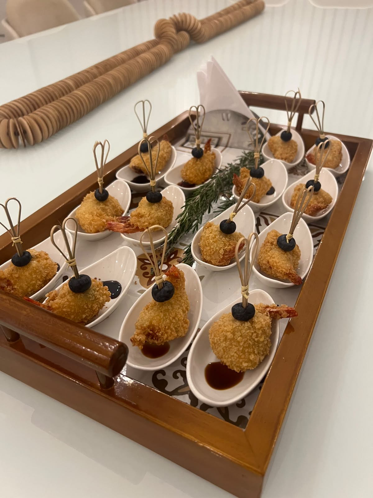 bandeja de madeira com colheres porcelanadas de camarão empanado com azeitona e molho, decoradas com alecrim, 