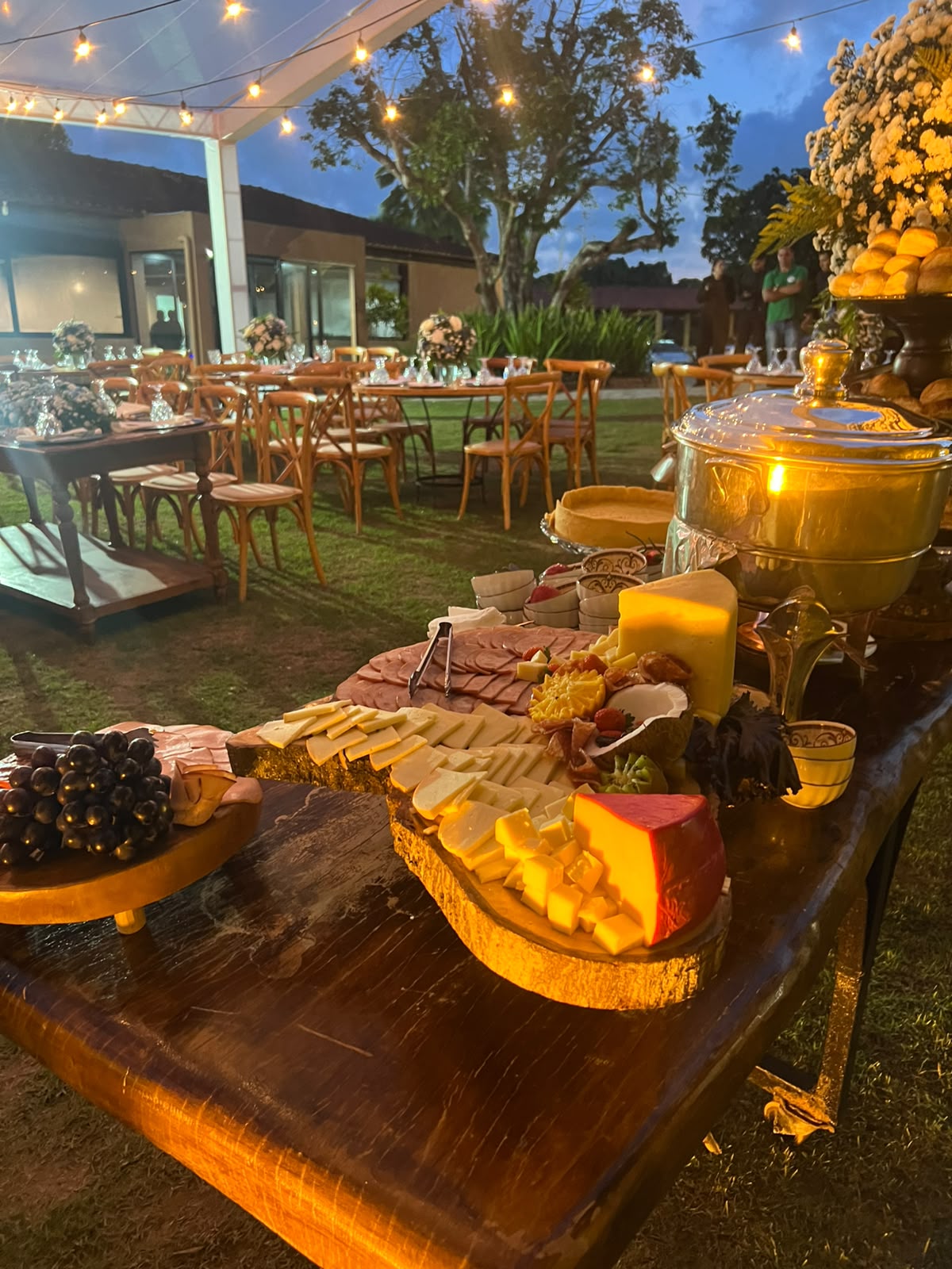mesa rústica de buffet com tábuas de queijos, frios, frutas e panela de fondue em evento ao ar livre com mesas