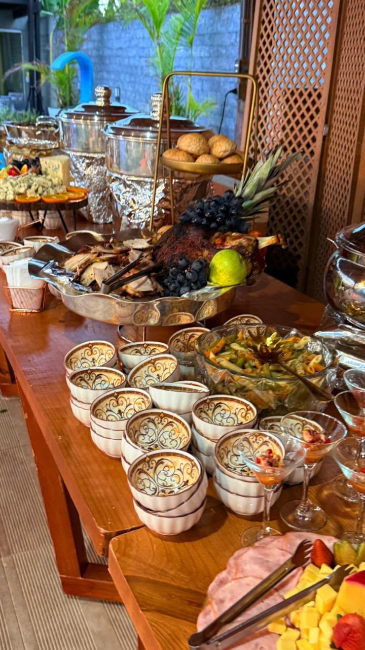 mesa de buffet de eventos com carnes, frutas, taças e potes de porcelana servidos em aparadores de madeira