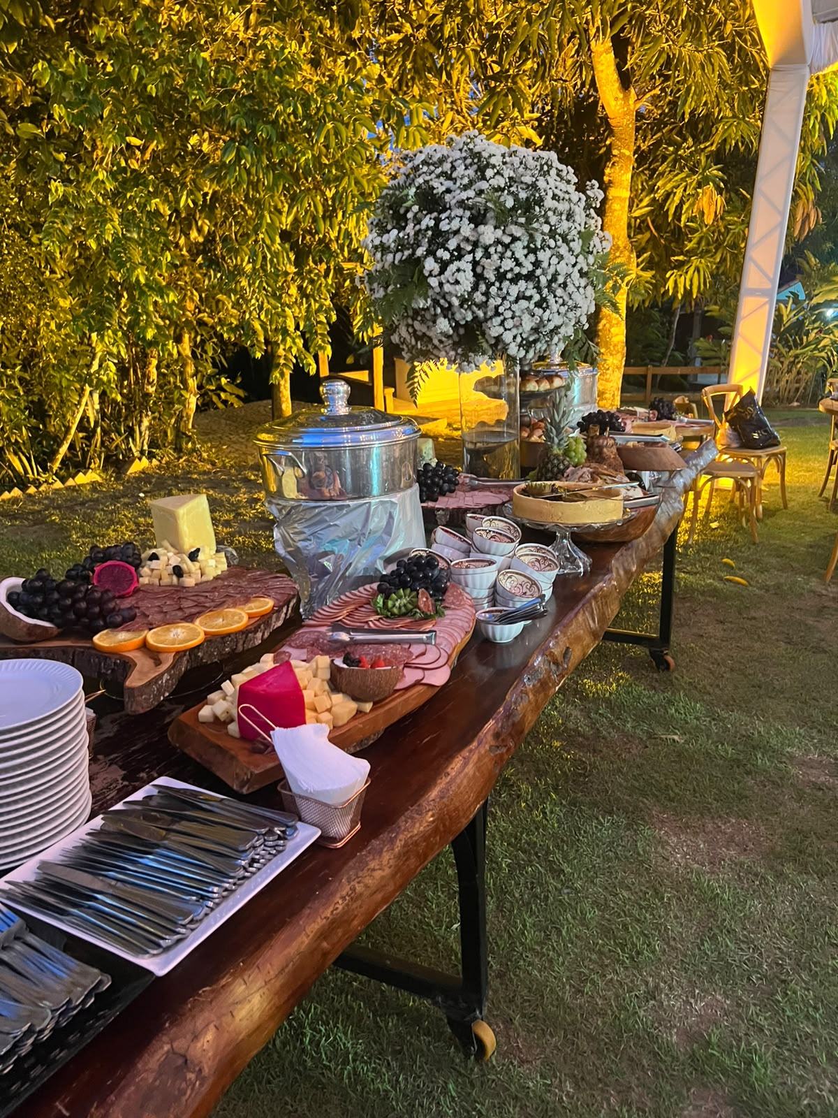 mesa rústica de buffet ao ar livre à noite com tábuas de frios, queijos, frutas, banquetas de prata e arranjo 