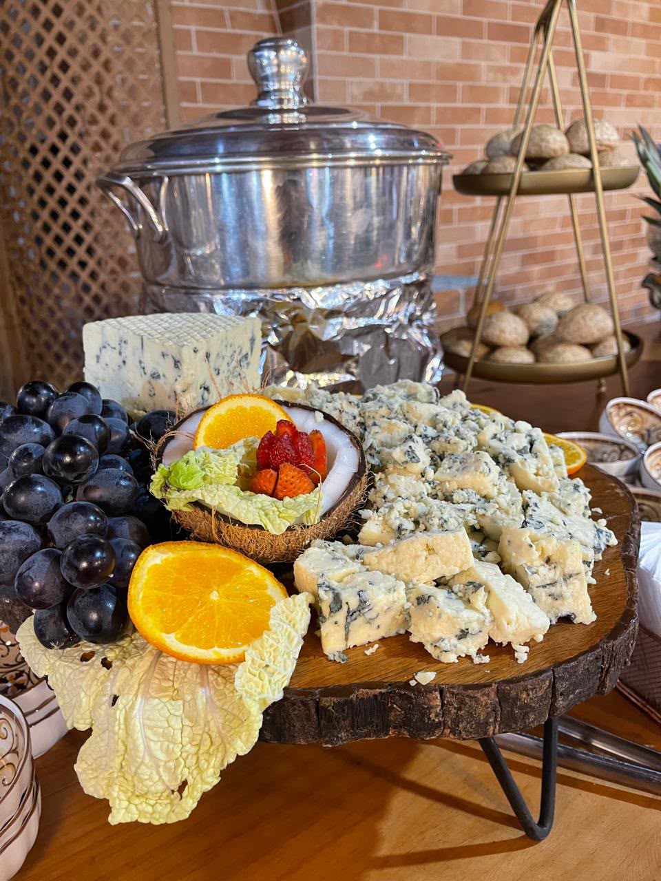 tábua de madeira com queijos gorgonzola, uvas, laranjas e morangos, com panela de inox e pães num buffet ao fu